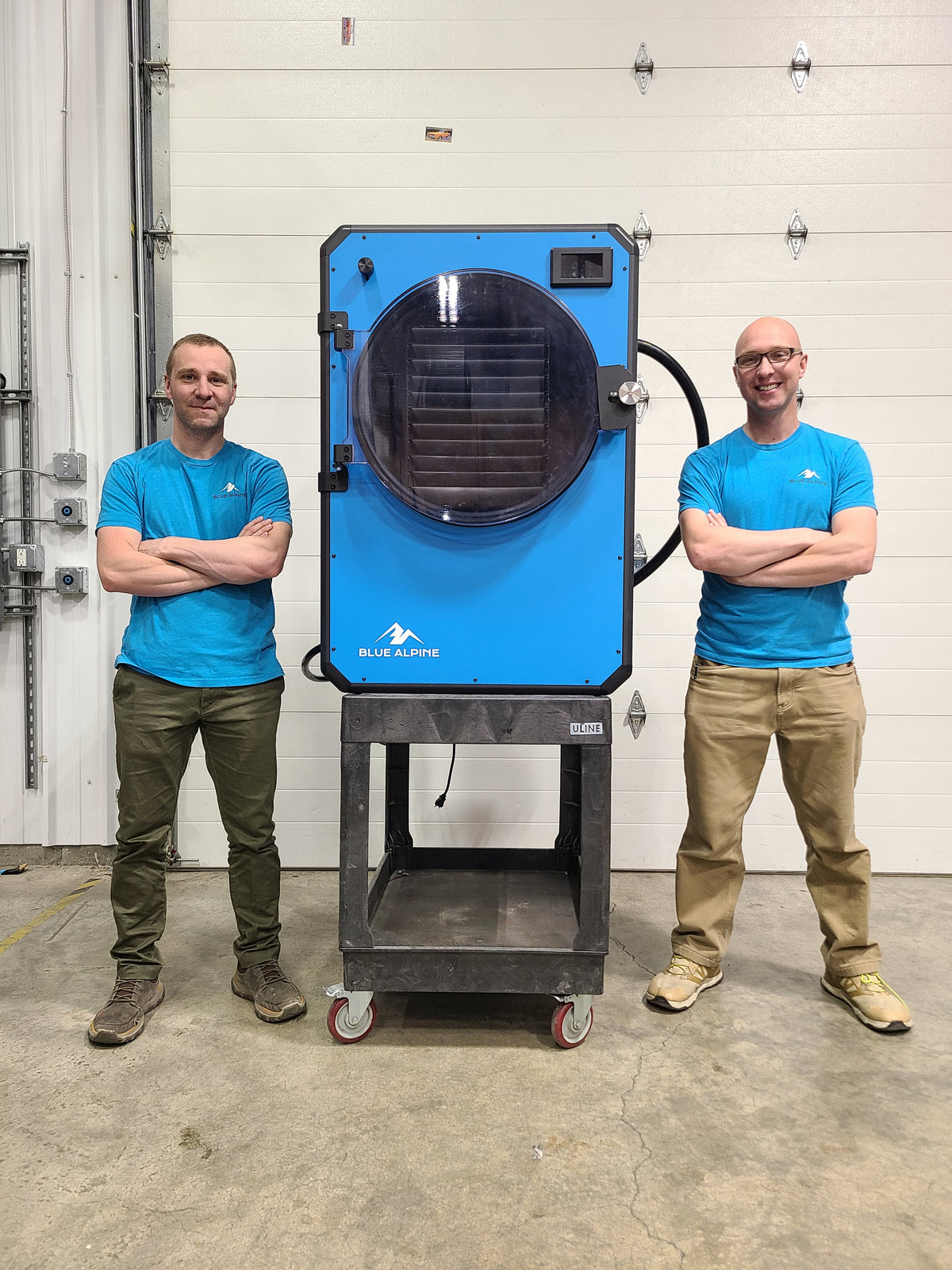 Two people standing next to a blue industrial dryer in an indoor setting.