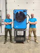 Two people standing next to a blue industrial dryer in an indoor setting.